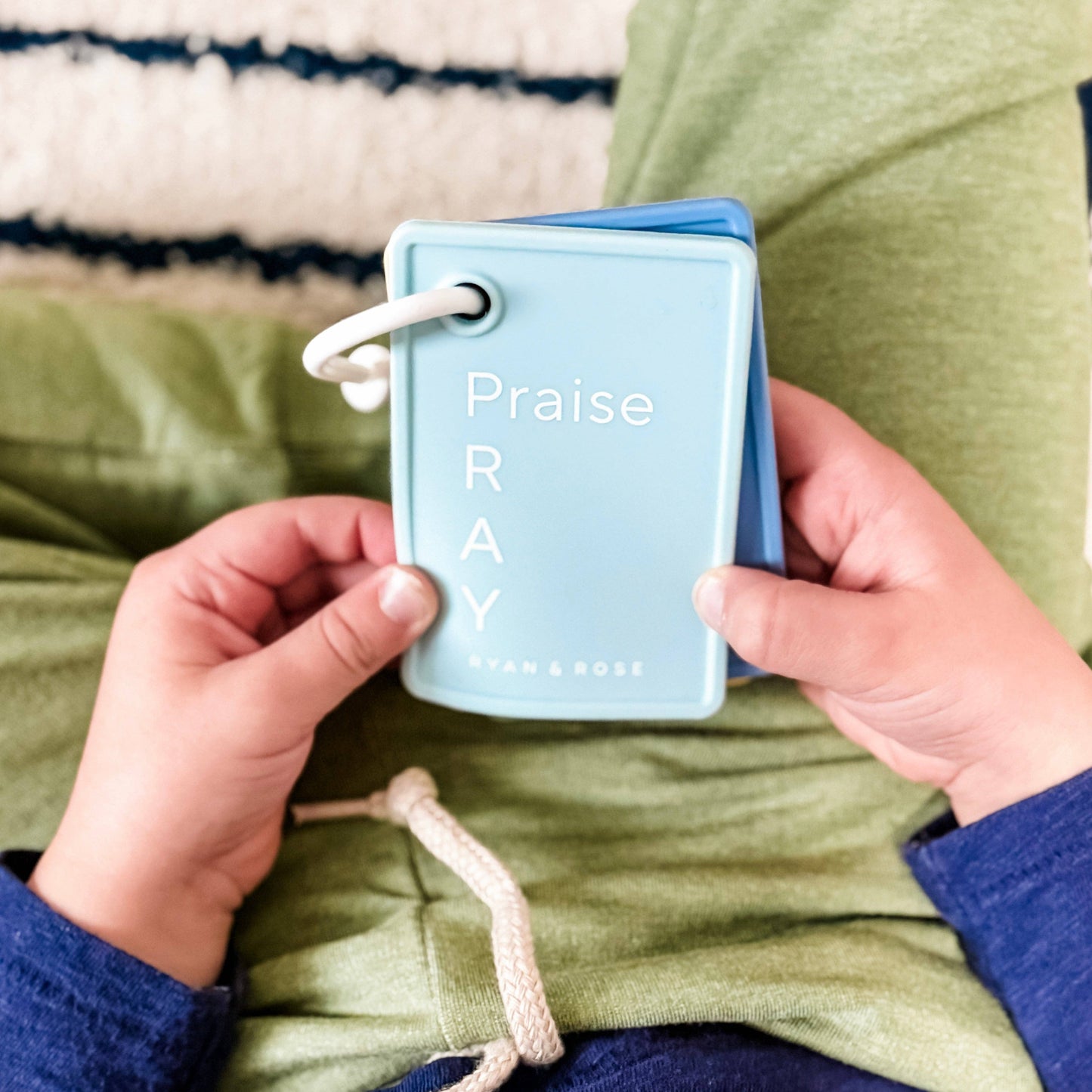 Person holding a small blue book titled 'Praise RAY' with a green jacket and striped fabric background.