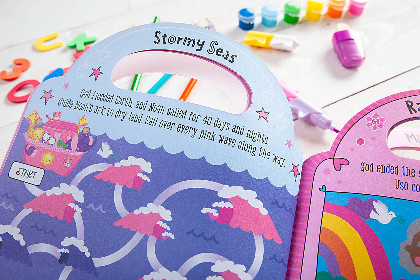 Children's pink activity books on a wooden surface with crayons and stickers.