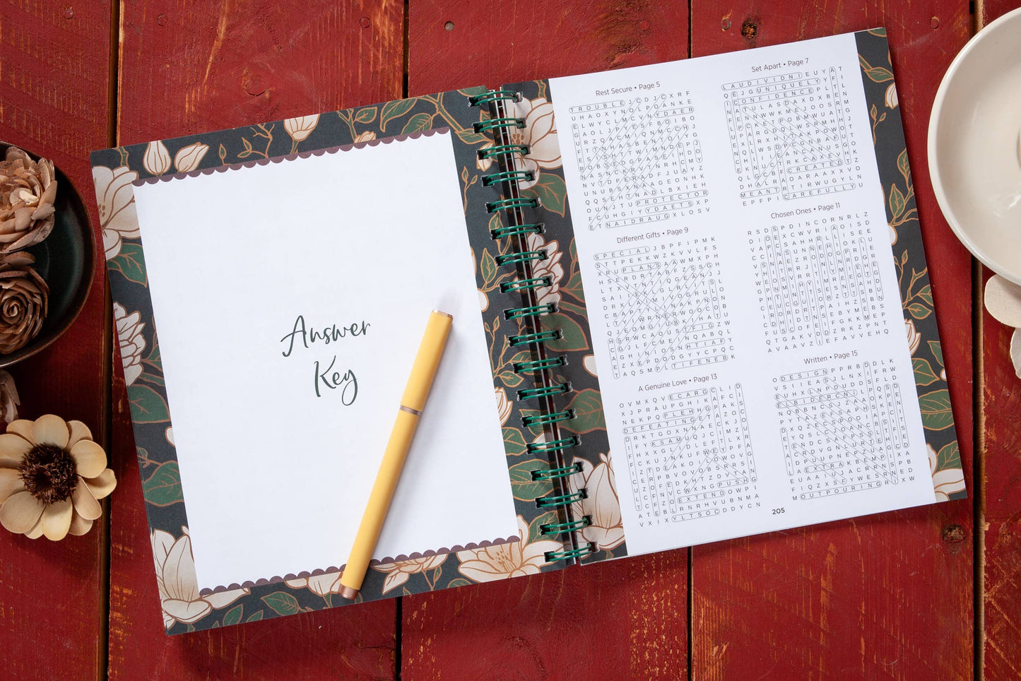Large print open floral devotional word search book on a table with  various objects surrounding the book.