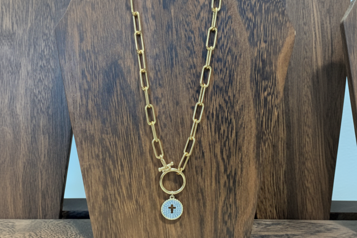 Gold chain necklace with a hollow cross pendant surrounded in intricate cubic zirconia stones placed on a wooden stand.