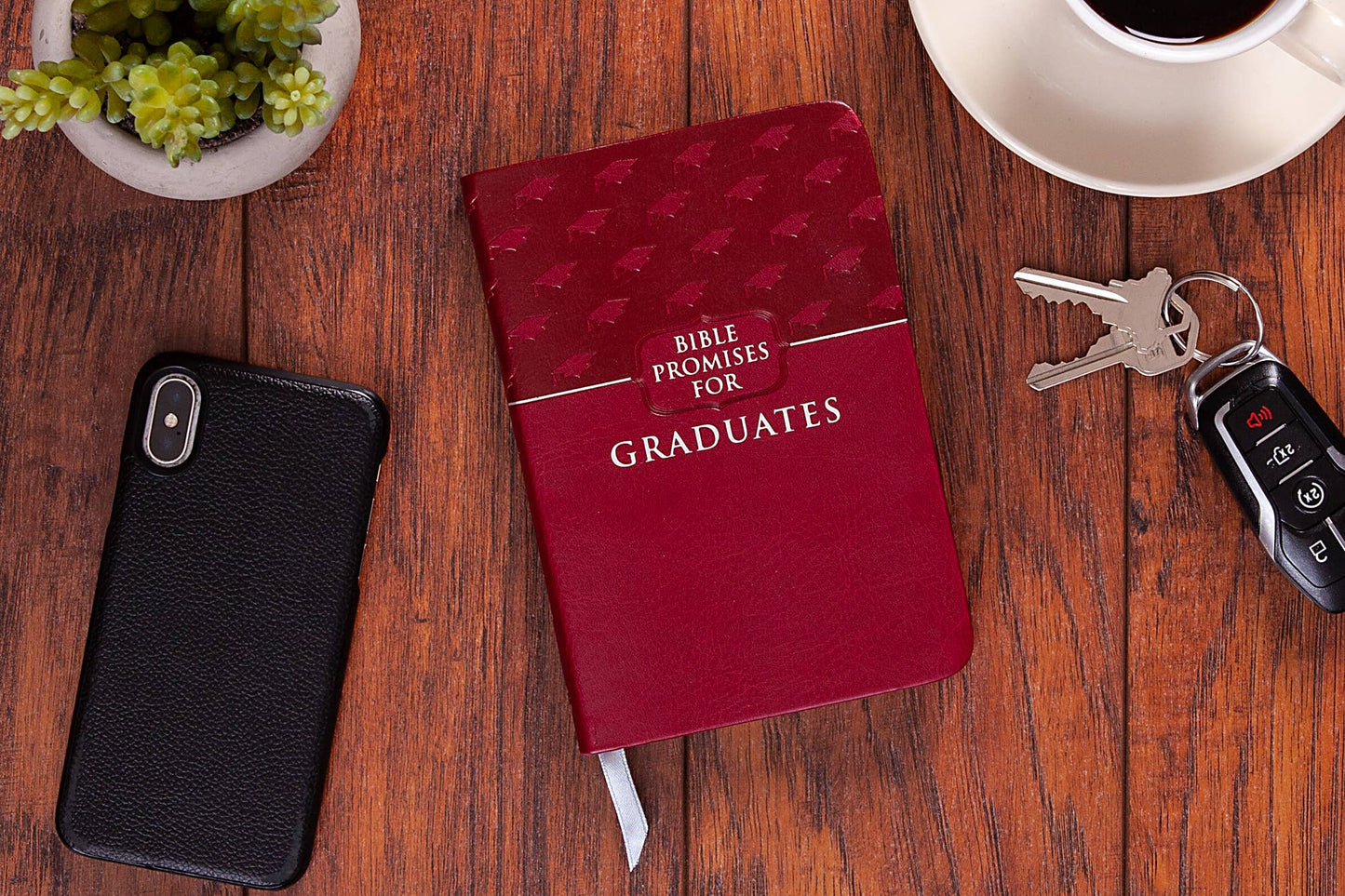 Red book of Bible promises for graduates sitting on a wood table with a phone, keys and plants around the book.