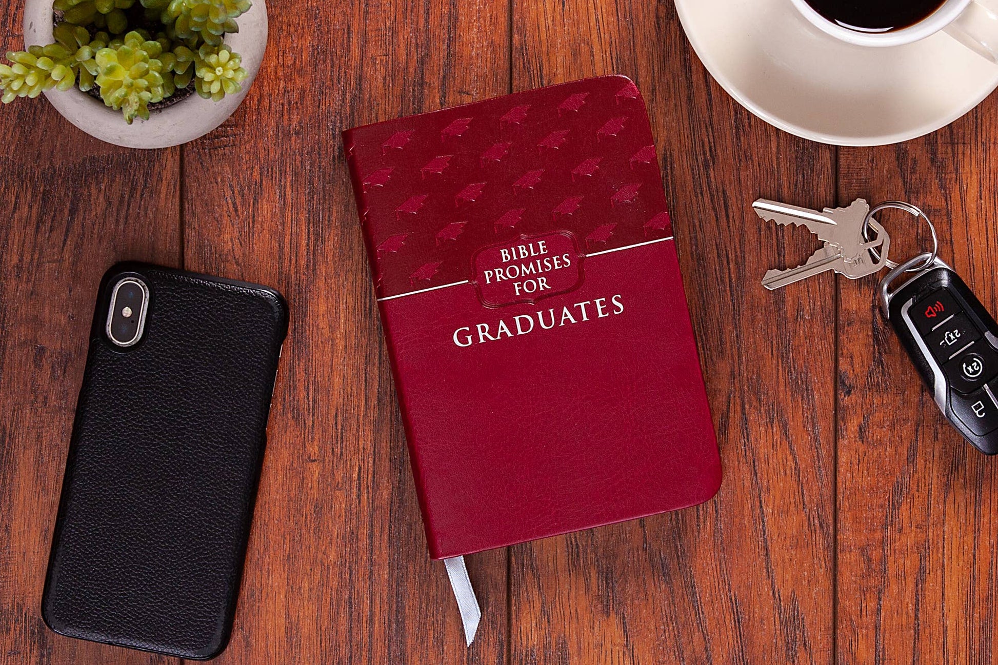 Red book of Bible promises for graduates sitting on a wood table with a phone, keys and plants around the book.