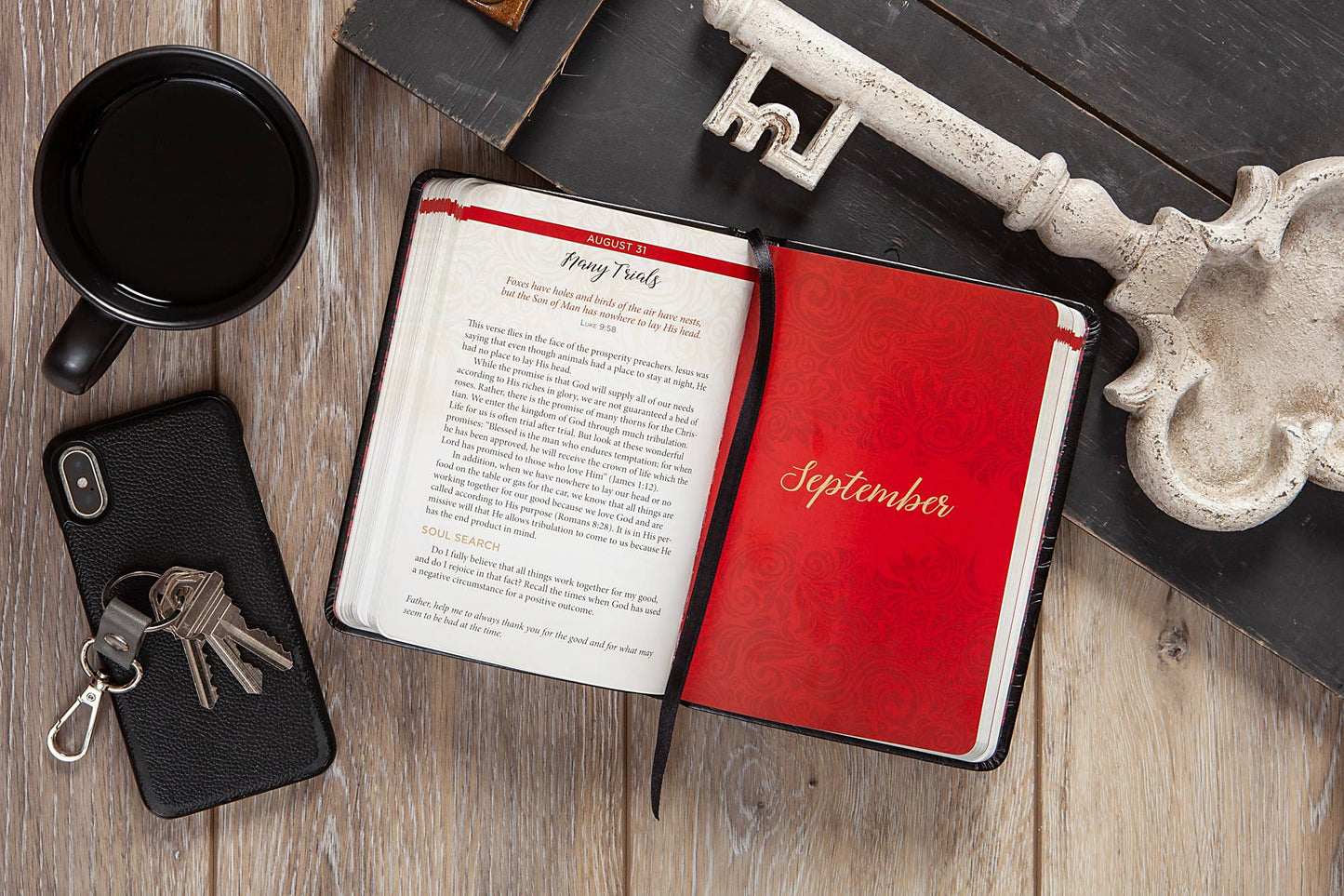 Open book with a red cover labeled 'September', black mug, phone, keys on a wooden surface.