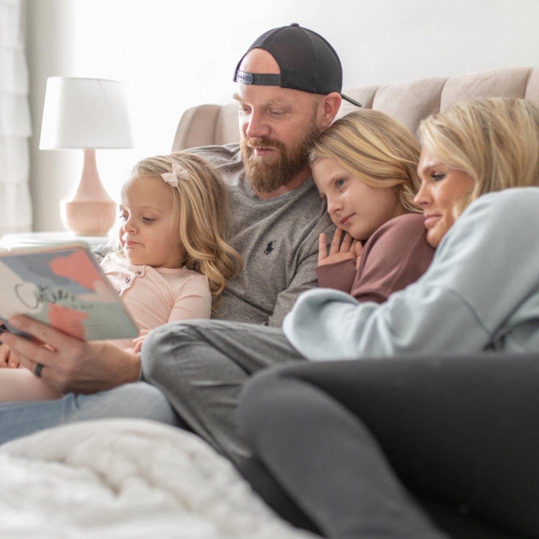 Family of four sitting on a couch, reading a book together in a cozy living room.