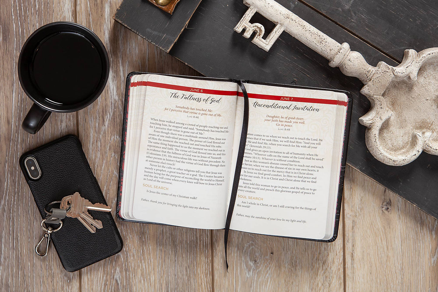 Open book with a red cover , black mug, phone, keys on a wooden surface.