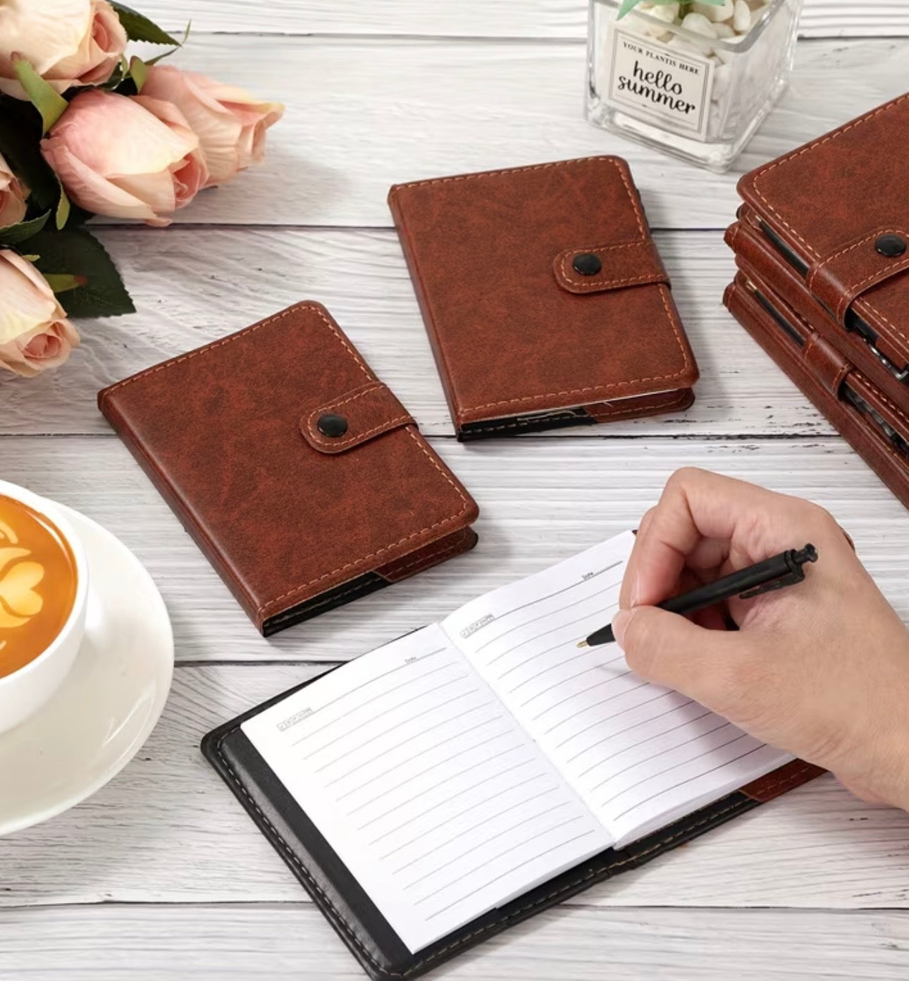 Brown vinyl mini notebooks with snap closures on a wooden surface with a hand writing in one of them, next to a cup of coffee and flowers.