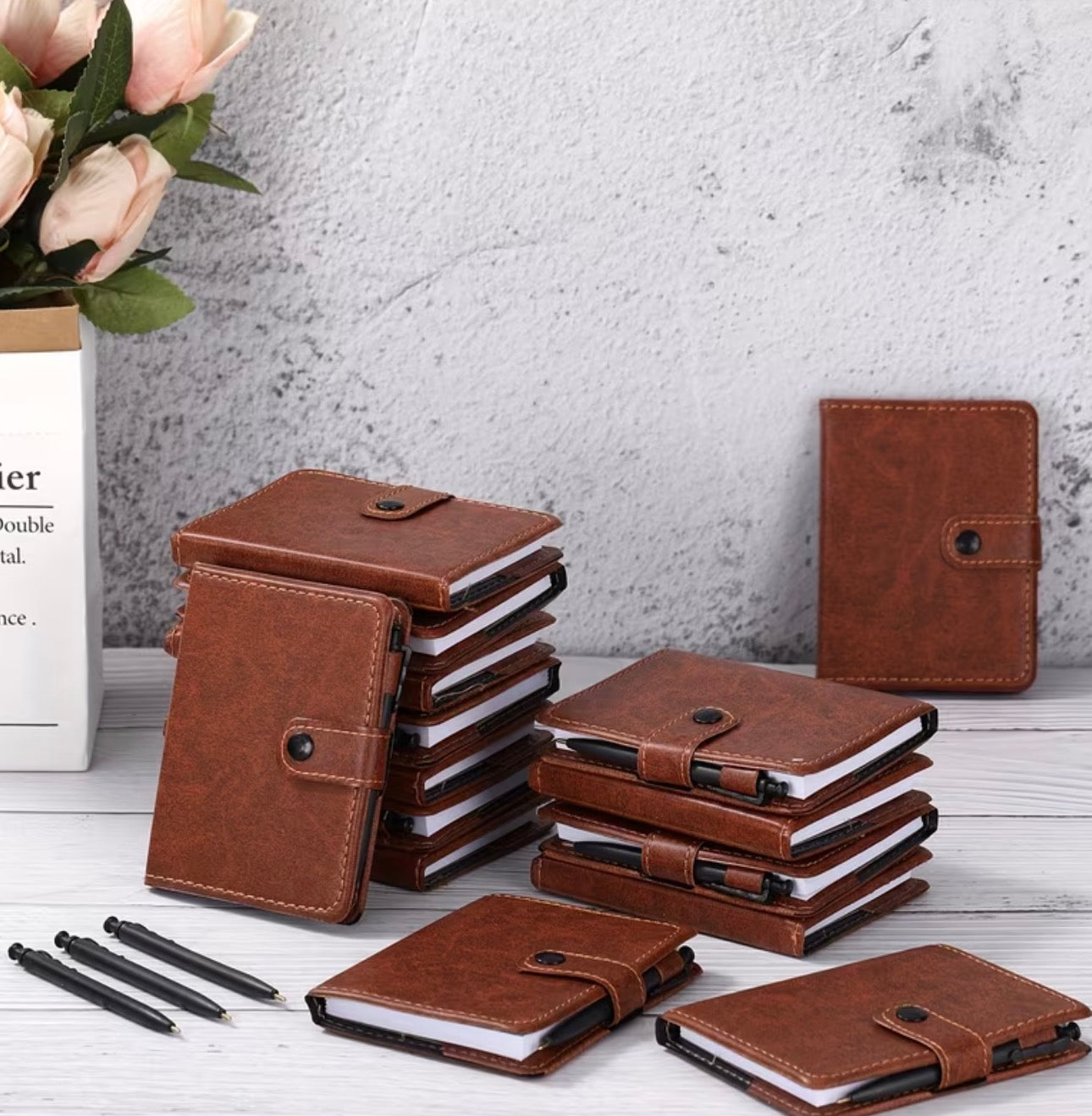 Stacks of brown vinyl mini notebooks with snap closures and pen holders on a white surface, with a gray textured wall in the background.