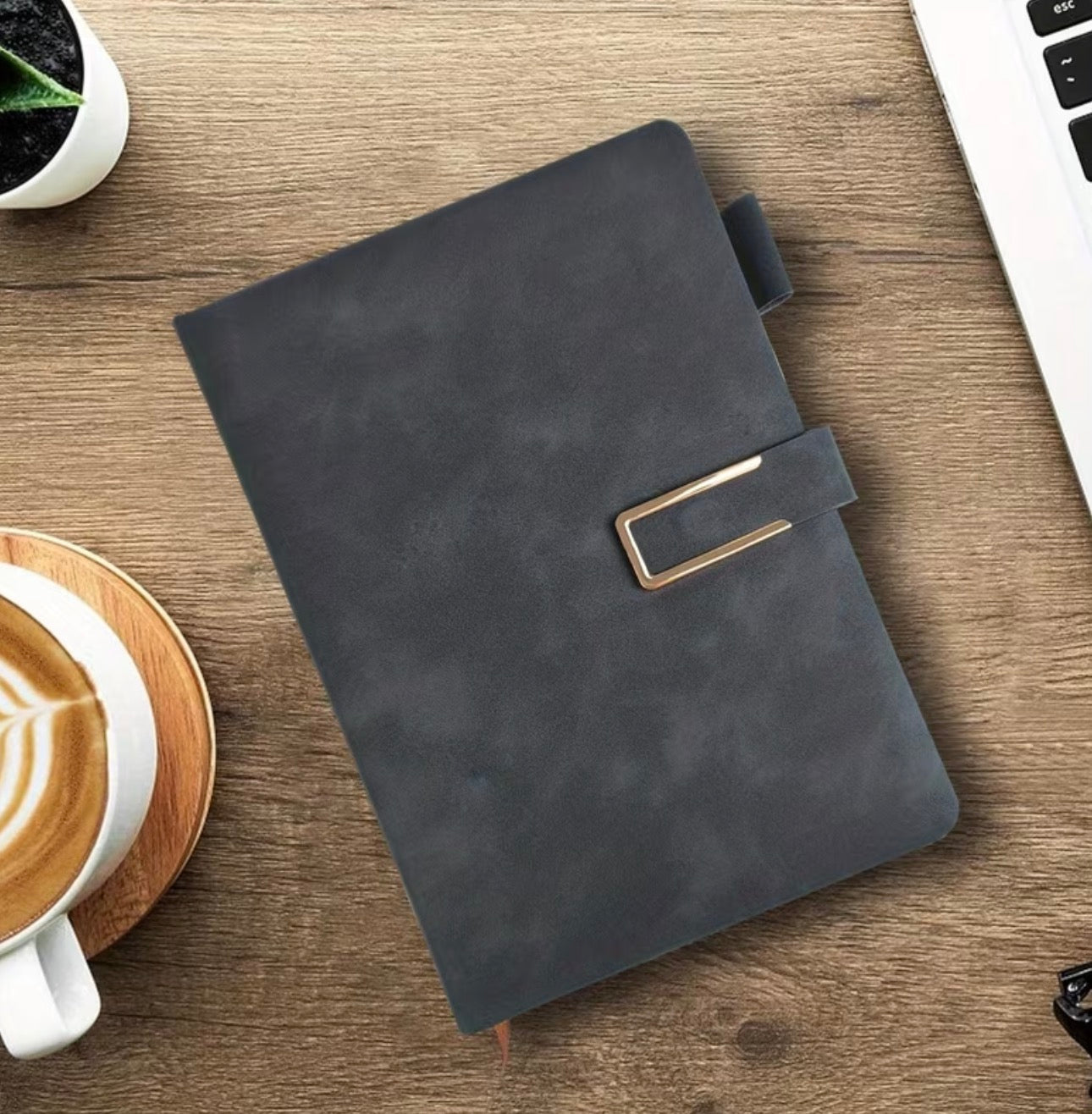 Black faux suede notebook with gold trimmed black magnetic closure buckle on a wooden desk with coffee and laptop.