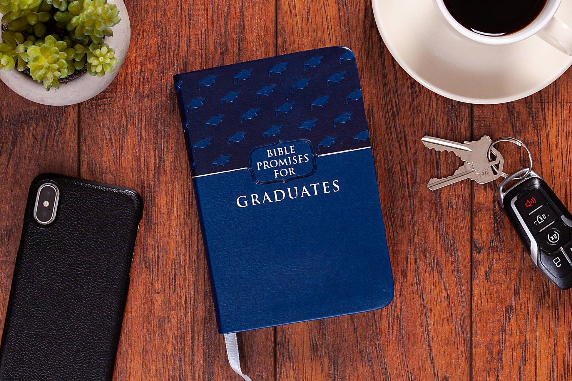 Closed book of Bible promises for graduates on a wooden table with a cup of coffee, keys, and a plant.