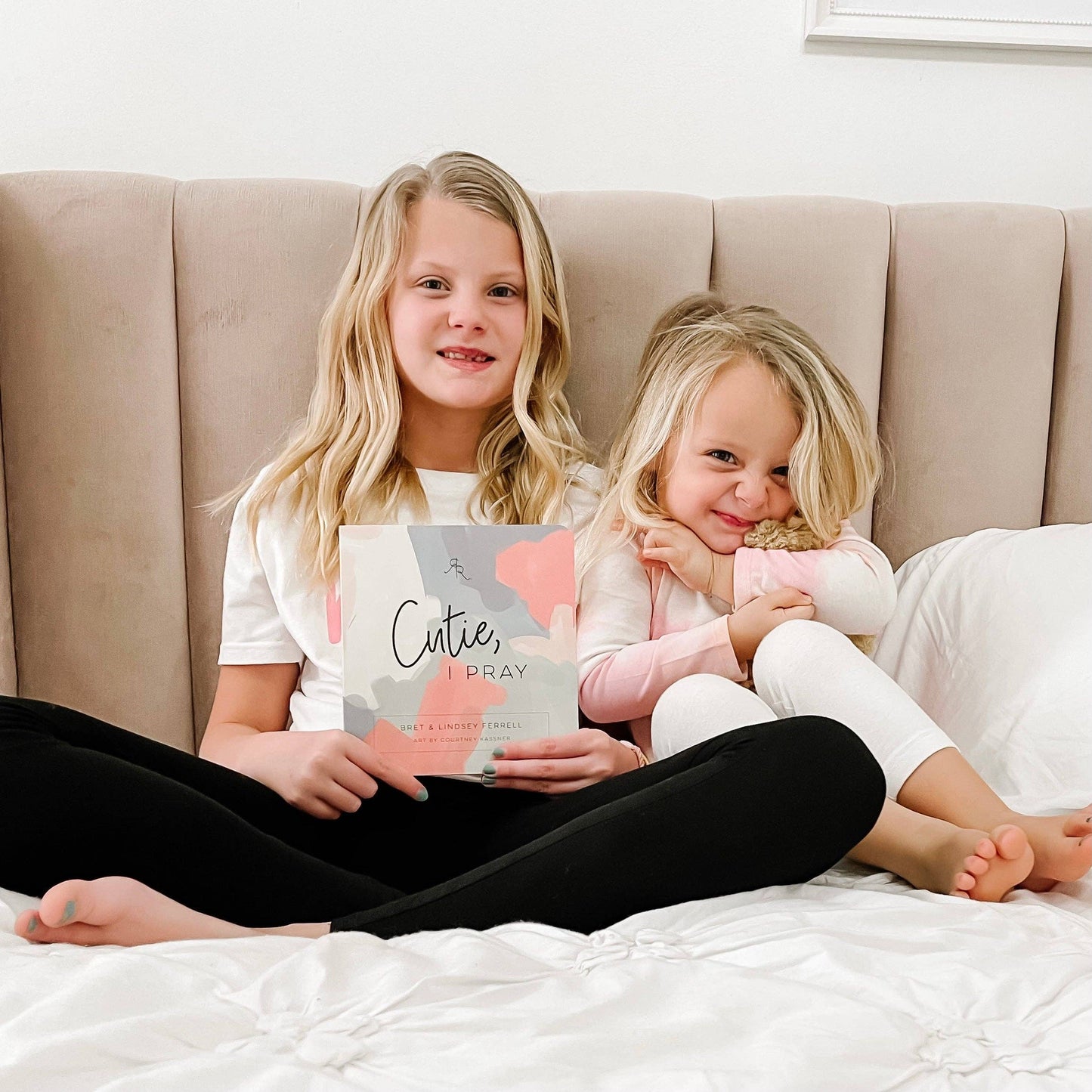 Two young girls sitting on a bed holding a book titled 'Cutie, I Pray'.