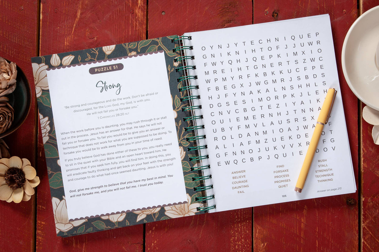 Large print open floral devotional word search book on a table with  various objects surrounding the book.