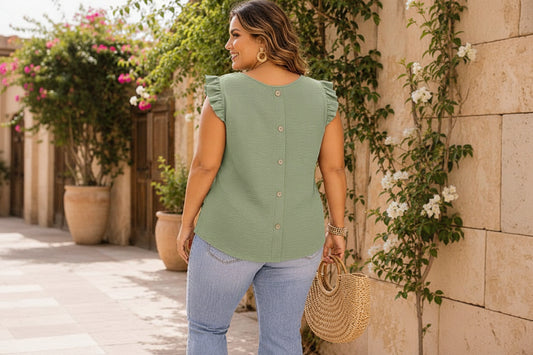 Woman wearing an sage top and blue jeans, holding a straw bag, standing outdoors.
